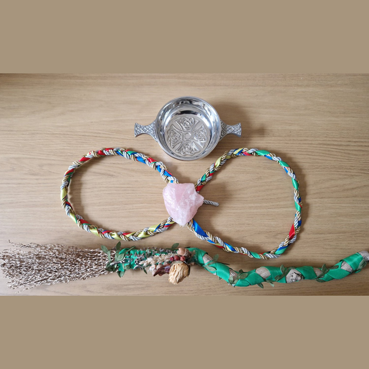 A silver Quaich with a Celtic engraving. Below this is a large rose quartz stone on top of a multi-coloured handfasting cord. Underneath this is a small broomstick which has been decorated with dried flowers and green ribbon.